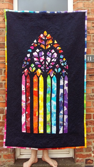 A picture of a quilt made to look like a gothic stained glass window being held up by someone whose bare feet are visible at the bottom. The background is the side of a redbrick house. 