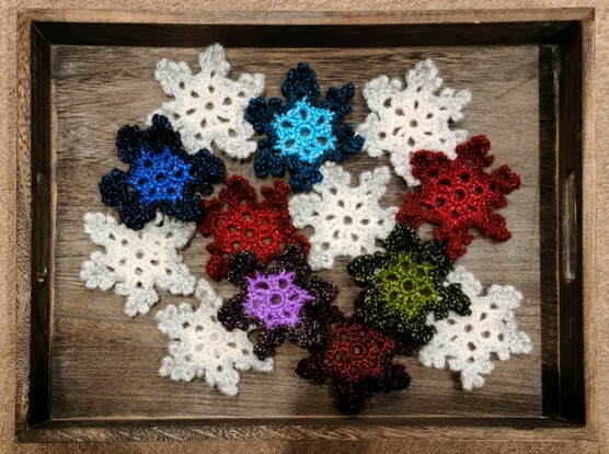 13 crochet snowflakes, six white, one indigo, one blue, one green, one purple, two red, one dark red.  Done in shiny metallic and sparkly yarn, about 5 inches across, light color centers and darker edges.  Displayed scattered in a gray wooden tray on a beige carpet.