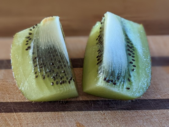 A kiwi fruit lies cut open on a wood cutting board, displaying its textured glistening green flesh and beautiful black seeds and white core.