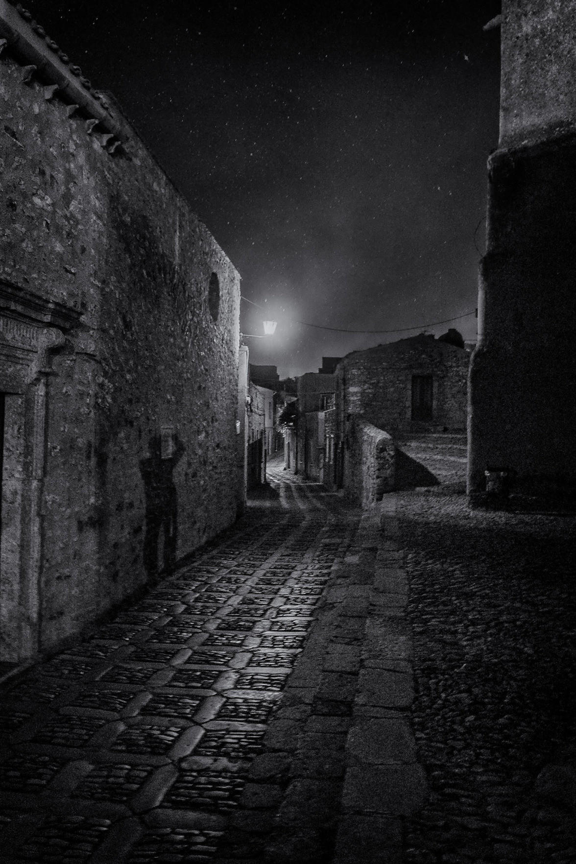 Dark cobblestone street in the Sicilian town of Erice with the photographer's shadow projected against the wall of a stone building.