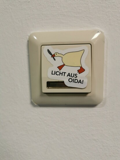 Ein Sticker auf einem Lichtschalter auf dem eine Gans mit Messer im Schnabel zu sehen ist darunter steht "Licht aus oida!"