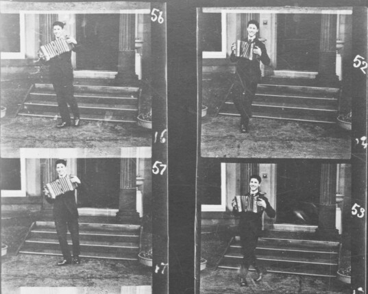 Four blown up frames of Le Prince's 1888 Accordion Player film showing much greater detail. Black and white images show Le Prince's son Adolph smiling while dancing side to side playing a small button accordion. Behind are the steps up to a house. The old film is degraded, but these four frames, are quite clear. There are hand written numbers beside the frames, 56, 16, 57, 17, 52, 13, 53.