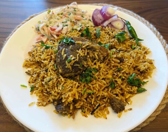 White plate filled with mutton biryani, raita, and raw onions and chillies.