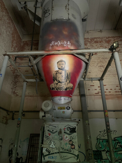 Photograph of industrial ruins covered in graffiti. Prominently featured on a metal chute is a well crafted stencil of a seated buddha, gold ringed with red, as if floating above a sea of scrawled tags.