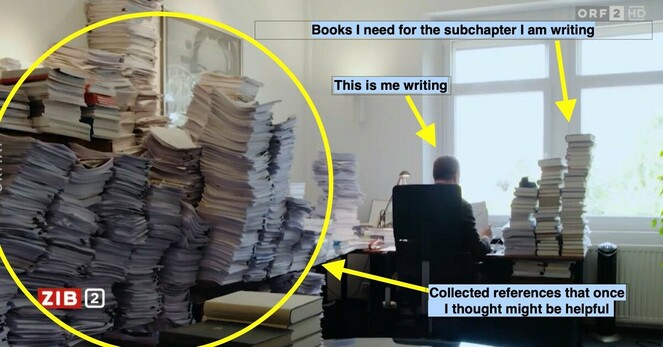 Photo of a working desk with loads of papers, and someone working at a laptop.