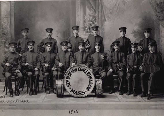 Antique group photo of the New Bedford, Massachusetts concertina band. 15 men in uniforms formally posing in two rows, one standing one sitting. With a bass drum in front with the name of the band. Two smaller drums are leaning against the bass drum. All the seated players have concertinas held on their knees.