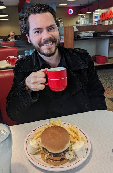 Bearded man at restaruant counter holding a cup of coffee over a double-deck hamburger.