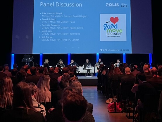 photo of panel of speakers at POLIS annual conference. in the background, the list of speakers:
- Elike van den Brandt (minister for mobility, brussels capital region)
- David Belliard (deputy mayor for mobility, paris)
- Carlotta Bonvinici (deputy mayor for mobility, reggio emilia)
- Janet Sanz (deputy mayor for mobility, barcelona)
- Seb Dance (deputy mayor for transport, london)