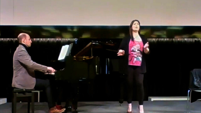 Blurry still frame from a Zoom video. Sitting on the left pianist Giacomo Marignani, wearing a gray jacket, dark pants and tan shoes. He is playing a grand piano with the lid open. In front of the piano opera soprano Megan Marie Hart, wearing a flowing black jacket over a pink top with a pop-art print, black tight pants and high-heeled shoes. Scene from GCHS 2021 Spring Chautauqua Virtual Concert Series.