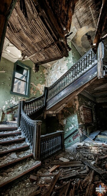 A view up a crooked wooden staircase. The walls are a mint green that is peeling away to reveal white underneath. Another stairwell leading down is mostly obscured. High above the first landing there is a window and a pail sits on one of the steps. To the right another hallway leads off into darkness