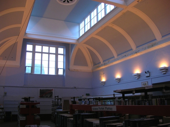 Interior of a tall rectangular library hall, painted in pale blue. Around the upper wall, deep curved shapes with bold white bracing lead to a windowed box set into the ceiling, at the centre of which is an ornate vent. Large windows let in daylight from one end. Along the length of the hall, electric lamps in sconces illuminate upwards.
At ground level, many bookshelves, some rather bare and forlorn.
(Alternative technical description from Historic England: "the ... library hall has a deep coffered ceiling with full-length lantern above, ceiling vents and a strainer arch with finials.")