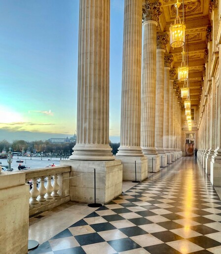 Loggia of the Hôtel de la Marine in Paris at twilight. There are black and white tiles on the floor and lamps lighting the narrow walkway and the beautiful ceiling. On the left Corinthian columns and a view on the Place de la Concorde and the Grand Palais in the background. 