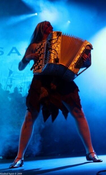 Netta Skog playing chromatic button accordion in the Finnish battle-metal band Turisas. She has long pale hair, red war-paint and is wearing ragged furs. Standing feet-planted on stage with blue smoke and lights behind her. 