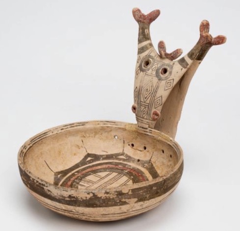 Pink-brown bowl with perforations near the handle, its base decorated with geometric patterns in brown and red. Handle includes a horned deer's head, its mouth resting in the bowl.