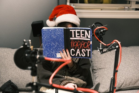girl wearing a santa hat and reading a book labeled "teen horror cast," sitting behind microphones.
