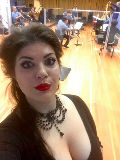 Selfie of a pale woman with unruly dark hair, wide open eyes with heavy spooky eye-makeup and bright red lipstick looking directly into the camera. In the background, orchestra members play bassoon, clarinets, violins and harp, and a conductor in casual attire. Soprano Megan Marie Hart and Maestro Giuseppe Finzi with the Staatsorchester Darmstadt in a musical rehearsals for Giacomo Puccini's opera La bohème on Halloween,  October 31, 2020.
