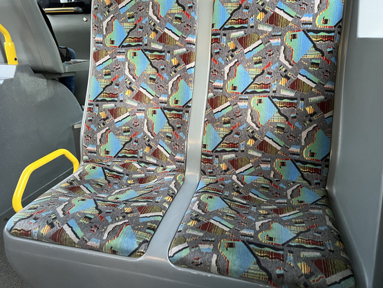 Sitzpolster in eim Bus mit einem Muster, das aussieht wie Scherben, in die bunter Müll gestreut wurde.