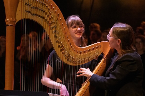 Een grote harp, rechts in beeld de harpjuf met hand op de snaren, achter de harp de leerling, lang meisje. Op achtergrond en door de snaren heen, zie je publiek. 
Juf en leerling kijken elkaar aan. Beiden in het zwart gekleed.