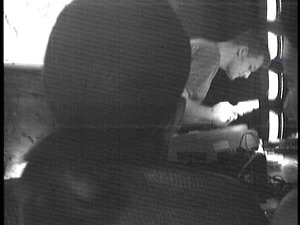 A black and white photo of Bitshifter performing at The Remote Lounge. He is hunched over his Game Boy and staring intensely at it. There is a big head in the foreground that blocks most of the image. The image looks like it was taken with a low-tech security camera. 