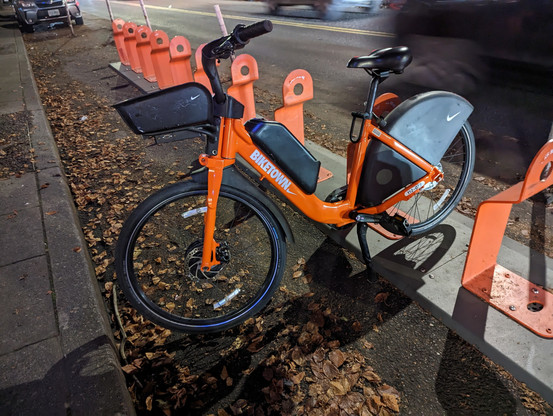 A new Biketown bicycle locked to a Biketown station rack. The dominate color of the bike frame is orange with black accents for the handlebars, seat, battery, and fenders. The bike is similar to the old style bike but more angular and uses a traditional bike chain instead of the shaft drive that the system launched with. This differs from the previous image in that the non-drive side of the bike.