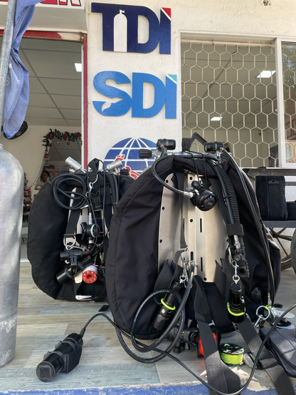 Dos equipos de buceo técnico de frente: alas, backplates, reguladores de dobles y carretes. Al fondo se ven los logos de SDI, TDI y PADI.