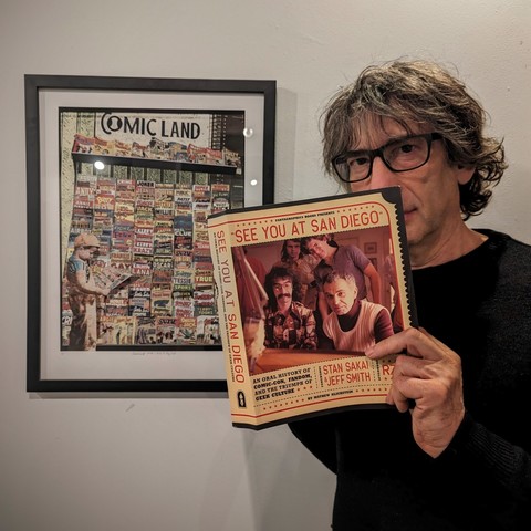 An elderly gentleman in need of a haircut standing in front of a 1940s photograph of a kid at a comics stall pretends to peruse a copy of See You At San Diego, an oral history of the San Diego Comic Con.
