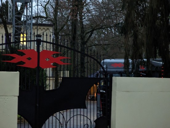 Ein Flügeltor vor einer Einfahrt auf dem das Triple X und das Batman Logo angebracht sind. Im Hintergrund sieht man ein Luxus-Camper mit Triple X Logo.