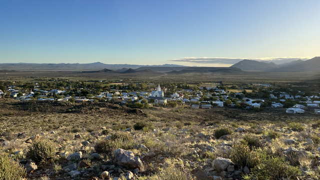 Wide view of the little town of Prince Albert in South Africa, with a white church spire at the centre, surrounded by whitewashed cottages and an expansive valley with fynbos shimmering in the light of sunrise. 