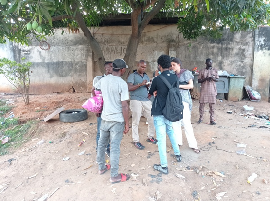 Unter einem Baum auf sandigem Boden stehen junge Männer im Kreis mit einer weißen Frau. Sie wirken ratlos. Teilweise haben sie Rucksäcke oder Beutel bei sich. Einer tippt in ein Handy. Drumherum liegt Müll.
