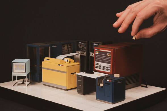 Miniature model of a 1950s mainframe computer including tape drives and printer. A hand is holding a miniaturized punch card the size of a thumbnail