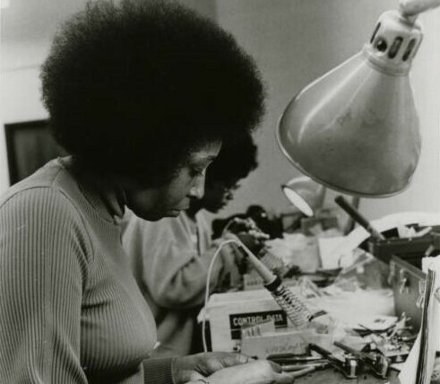 This black and white photo is of two African American women standing at assembly tables doing assembly work with microcircuitry. In the image is a lamp, soldering tool, and various components.