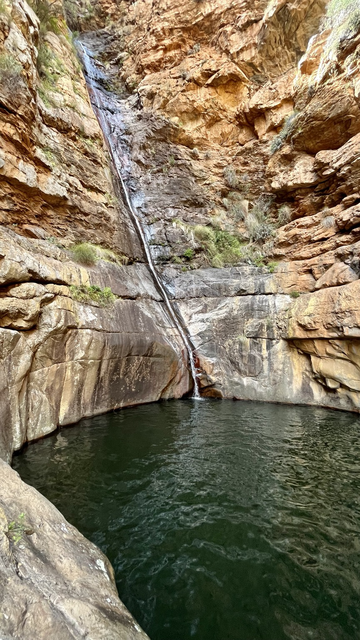 A slim, straight waterfall runs over smooth rocks into a dark mountain pool