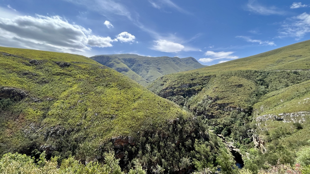 Wide view of lush green mountains, a dark ravine and partly cloudy sky. 