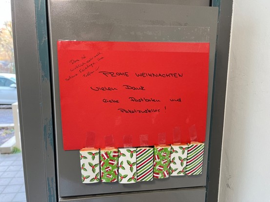 Zettel im Hausflur mit Glückwünschen an die Postboten. Darunter angeklebt: Schokoladentäfelchen. Die Postfrau bedankt sich artig in der Ecke des Zettels. 
