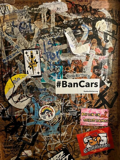 White #BanCars sticker on a wall covered with graffiti and other stickers.