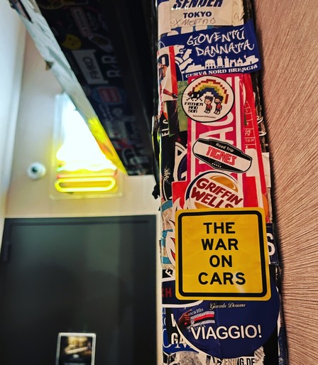 Yellow "The War on Cars" logo sticker on a post with lots of other stickers. A neon sign is out of focus in the background.