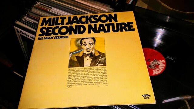 Front cover of "Milt Jackson – Second Nature: The Savoy Sessions" with big black letters on a creamy cover, a drawing of Milt looking surprised in the middle. Behind it plays the record, half the red Savoy label on the right, a box of records on the left.
