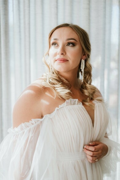 Bride in her dress backlit by white curtains 