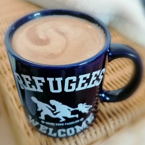 Foto: Ein Becher voll Milchkaffee, blau, Beschriftung weiß: Refugees welcome.