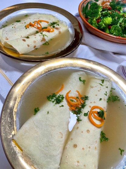 Two bowls of Crespelle In Brodo: rolled crepes in broth in clear bowls with a gold band. The crepes are garnished with carrot curls and minced parsley. There is a bowl of broccoli in the upper right corner.