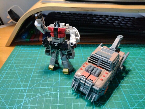 Photo. Transformers toys on a desk. Core Class Sludge in robot mode and Deluxe Scraphook in vehicle mode.