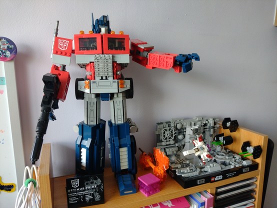 Photo. The Lego Optimus Prime model in robot mode on a shelf pointing with his left hand. Next to him is the Lego Death Star Trench model.