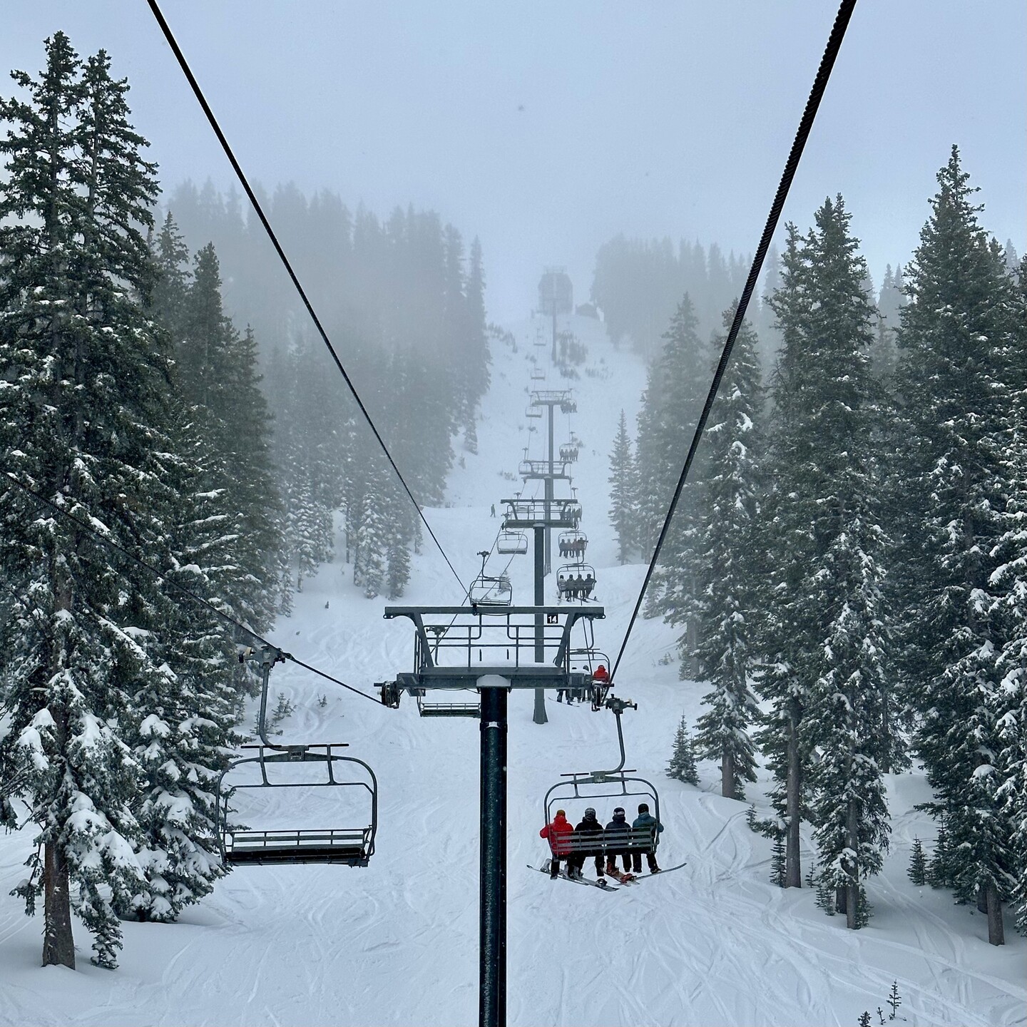 Photo from a chairlift looking up a mountain toward the top. Cloudy and snowy conditions with snow covered trees on both sides.