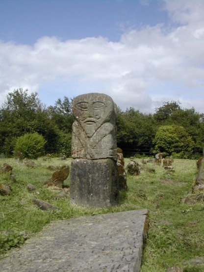 A large, grey, flat, stone slab lies at an angle in the left foreground. Behind it is a small (less than one metre talk) upright stone, carved with a face in the top third. You can clearly make out eyes, nose, what might be a curly moustache, mouth and a pointy chin.

The middle third of the stone is carved to show simple crossed arms. There is a horizontal crack between the middle and bottom layers.

The idol stands in a scrubby old graveyard with low headstones and an overgrown hedge in the background.