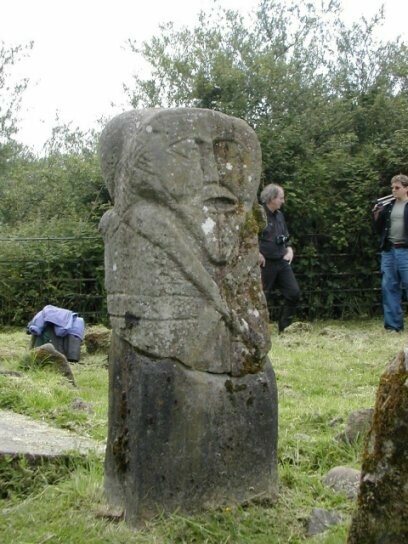 The rear of the small standing stone shows similar carvings to the front. This photo is taken from a slightly side angle so you can see the rear and the side of the figure. There are carvings at the side of the heads that might be hair or a pattern.

In the rear of the image is a hedge, with a couple of people standing beside it. One has a camera tripod over their shoulder.