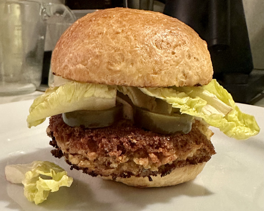 A Hoppin’ John Burger (vegan black eye pea plus veggies and nuts mixture) on a homemade potato bun. Pickled jalapeño slices, chutney mayo, and butter lettuce on the burger. 