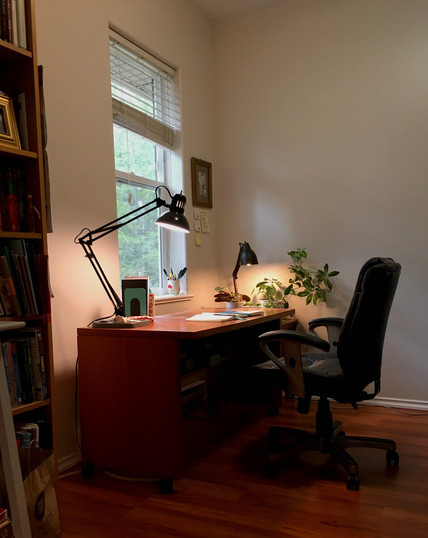 Photo of a small low wooden business-type desk pushed against a wall, beneath a small window, with a cushy black office chair.  The desk top is mostly empty except for an open day timer. Two black desk lamps on opposite corners of the desk. One casts an orange light on the desk, the other on some houseplants. 