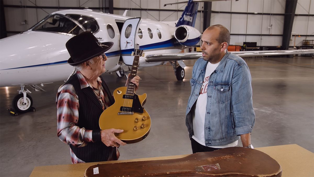 The guitarist is holding for presentation, a gold Les Paul in front of another man wearing a blue jean jacket. They are standing behind a table with a guitar case and are in an airplane hangar with a Lear Jet behind them.