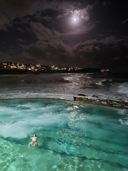 Swimmer breast stroking in fore ground  towards the other side of ocean pool lit turquoise, large breaking waves in sea outside pool, washing into pool, clouds scuddimg overhead lit by bright moon with city lights in background