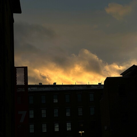 View of the mill in Maynard, Massachusetts silhouetted against a yellowy sunset. The color of the sunset is torn apart by unsettled clouds.